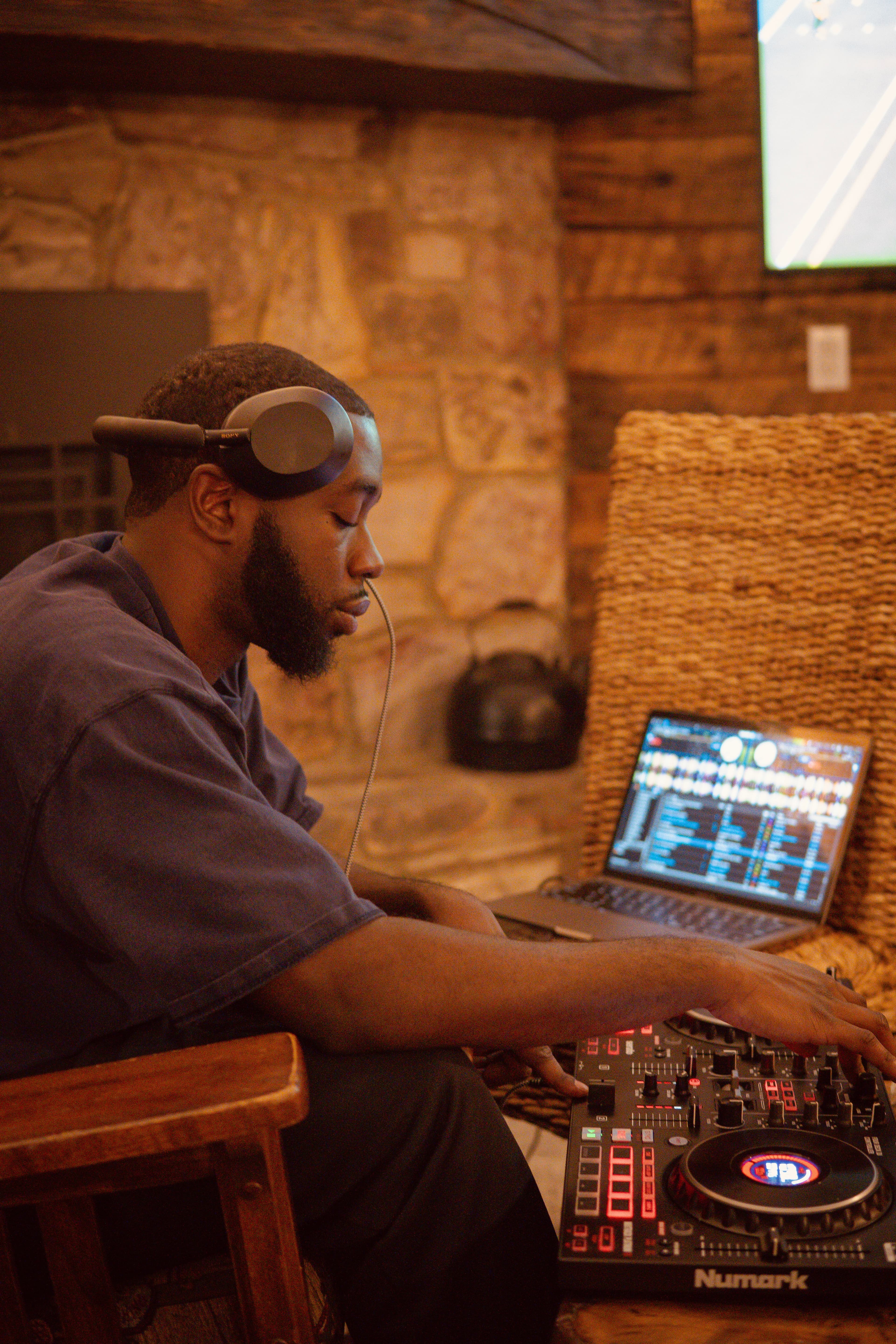 Mo wearing headphones, eyes down, working a DJ controller beside a laptop in front of a stone wall.