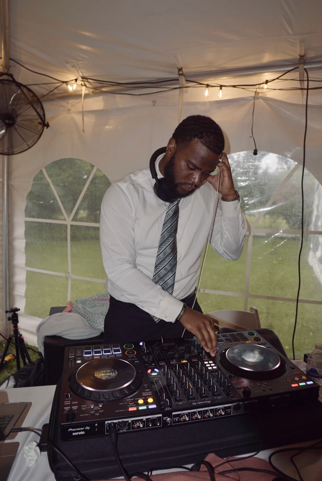 Mel in a white shirt and tie, headphones around the neck, mixing on a DJ controller inside an event tent.